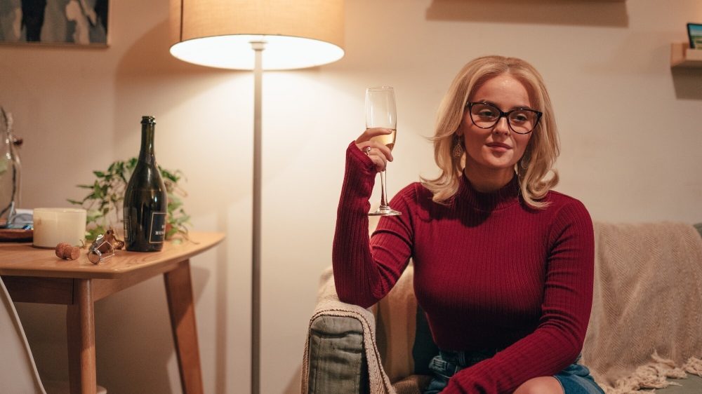 Millie Gibson as Ruby Sunday, a white woman with shoulder-length blonde hair and dark-rimmed glasses, wearing a long-sleeved red, formfitting top and holding up a wine glass with white wine.