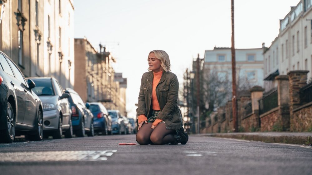 Millie Gibson as Ruby Sunday, a white woman with shoulder-length blonde hair kneeling in the street and looking distraught. She's wearing an orange long-sleeve shirt, jean skirt, black tights, and olive-green jacket.