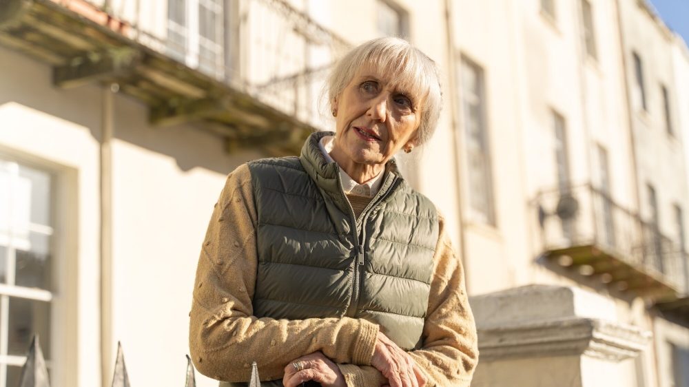 Anita Dobson as Mrs. Flood, an older white woman with a white-haired bob, cream long-sleeve shirt, and green puffer vest. She's looking past the camera with her head tilted.