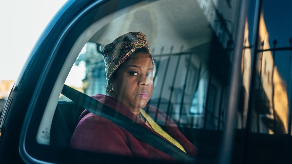 Michelle Greenridge as Carla Sunday, a black woman who looks tense in the backseat of a car. She's wearing a multi-colored headscarf, maroon jacket, and green top, and is buckled in.