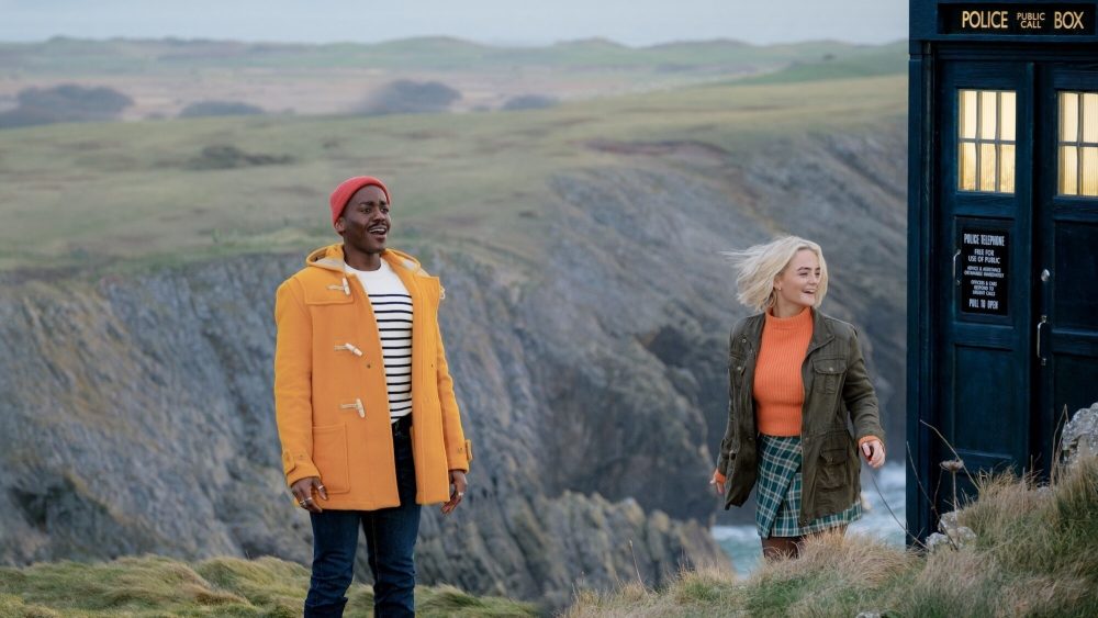 Ncuti Gatwa as the Doctor and Millie Gibson as Ruby Sunday. Ncuti is a black man in a red cap, bright orange jacket, white black-striped top, and blue jeans. He looks excited, as does Millie, a white woman with shoulder-length blonde hair, wearing an orange shirt, olive jacket, and blue-checkered kilt.