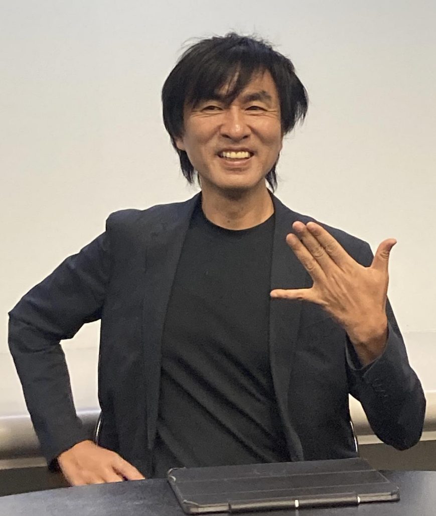 Kawamori Shoji. Smiling man wearing black jacket and undershirt holds out the little finger and thumb of his left hand, forming a flat symbol.