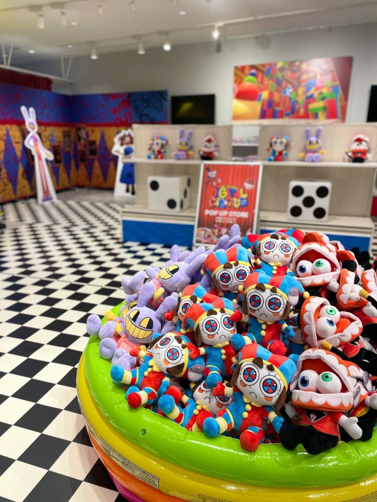 Pictures from The Amazing Digital Circus pop-up store in Japan. We see a bunch of Pomni and Cain plush toys in the lower right corner, with character cutouts on the back in a checkered floor pattern.