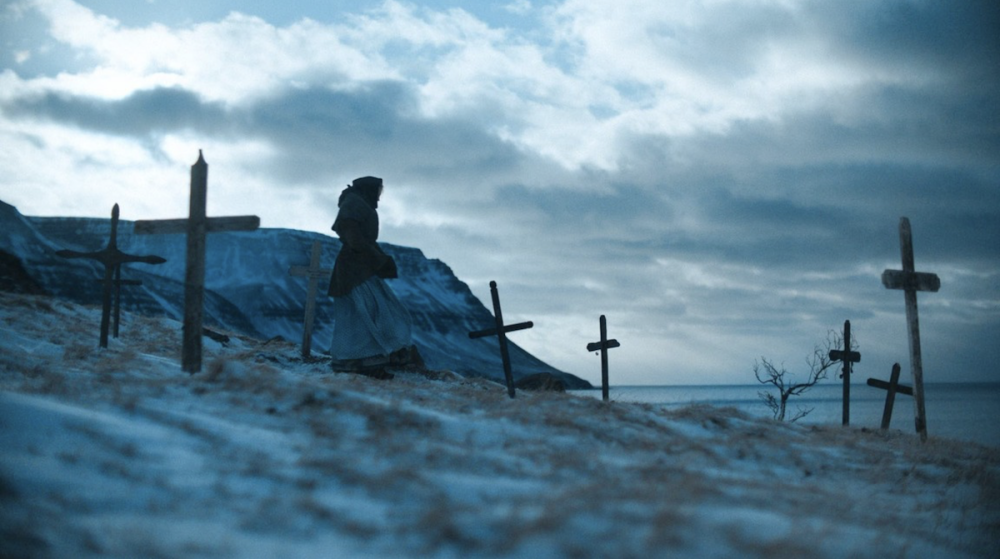 A woman walks along a gray shoreline dotted with graves. 